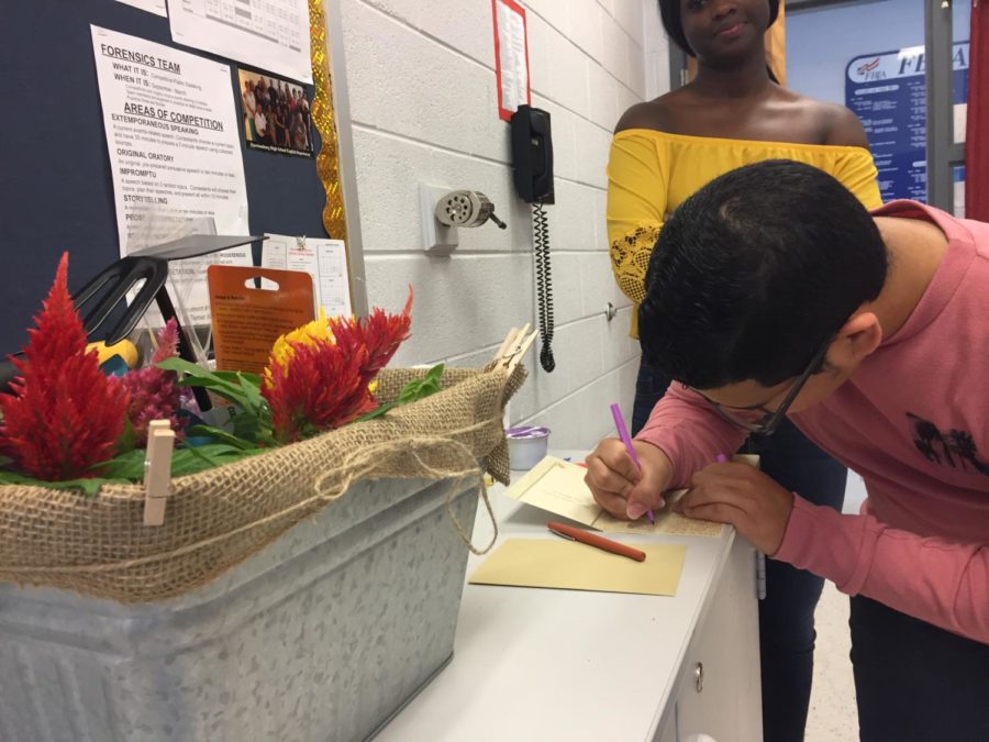 Junior Fernando Escobar-Medina writes the SCA thank you note to Cynthia Prieto for her retirement. The gift is on the left side of the screen.