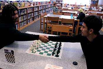 Two students shaking hands in the library, prepared to play a game of chess.