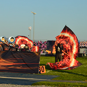 Girls in black outfits wave flags with the image of a streak of fire on them. The band is to the left, and an audience is in the back.