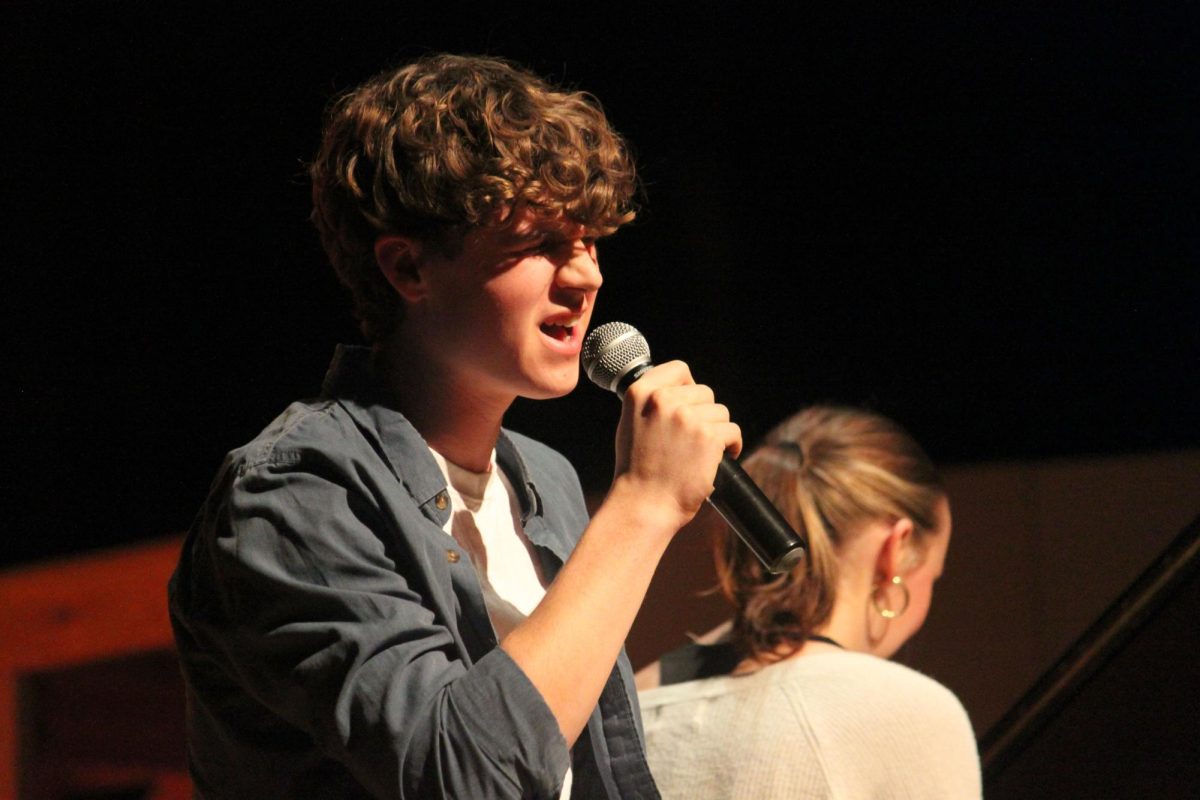 A boy holds a microphone with his mouth open while a girl sitting behind him is looking down at a piano that is not shown in the photo.