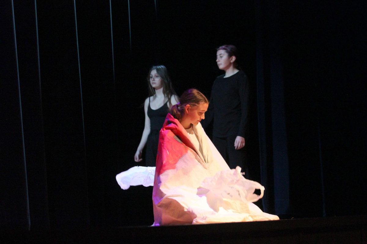 A girl sits on a stage wrapped in a tapestry holding plastic bags while two dancers walk by in the back