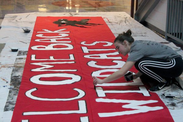 a girl squats while painting a black border on the words on a red banner