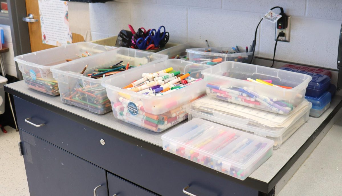 Art supplies are laid out on a wooden desk, with colorful paints, brushes, and pencils ready to go. Each tool has a unique history from past projects and creative times. Every student or teacher picking up a supply holds the power to bring another vision to life.